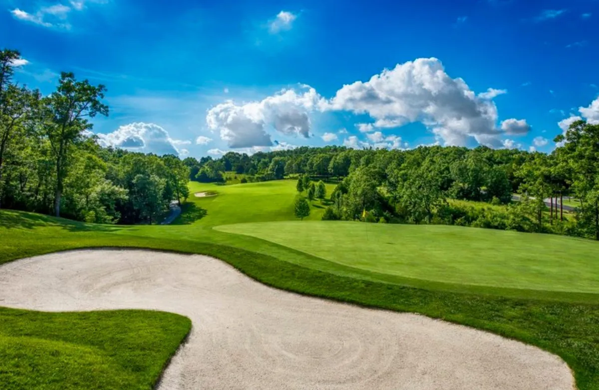 This image captures a lush, green golf fairway with a sand trap in the foreground, distant trees framing the scene, and a vivid blue sky overhead. The expansive view highlights the course's well-maintained grounds and the natural beauty of the Ozarks backdrop.