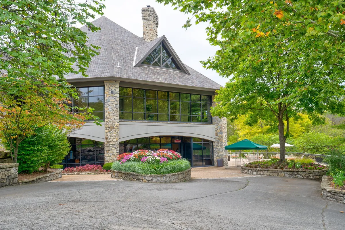 A scenic exterior with large glass windows, stone facade, colorful flower beds, and surrounding trees—inviting guests to explore nearby nature paths and enjoy outdoor spaces.