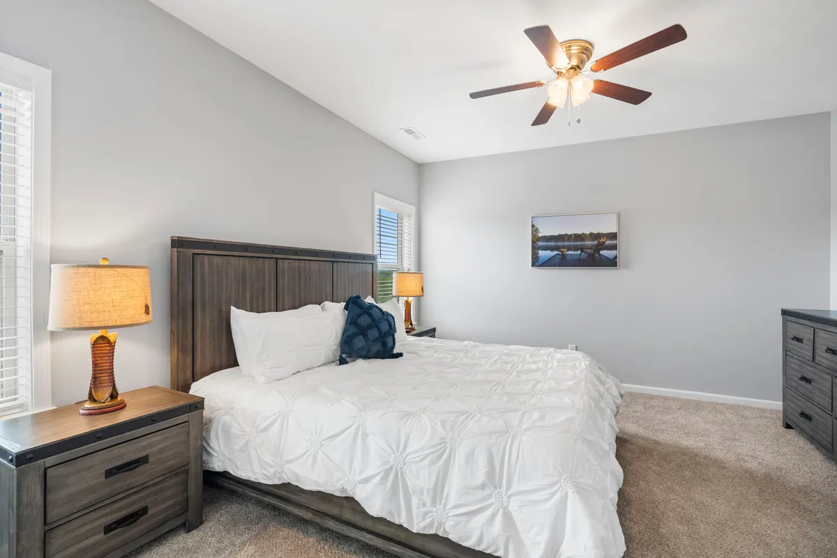 A tranquil bedroom featuring a large bed with crisp linens and plush pillows, flanked by nightstands and softly glowing lamps. Above the bed hangs tasteful artwork, while a ceiling fan gently stirs the air. The atmosphere is peaceful and inviting, perfect for restful sleep or quiet reading.