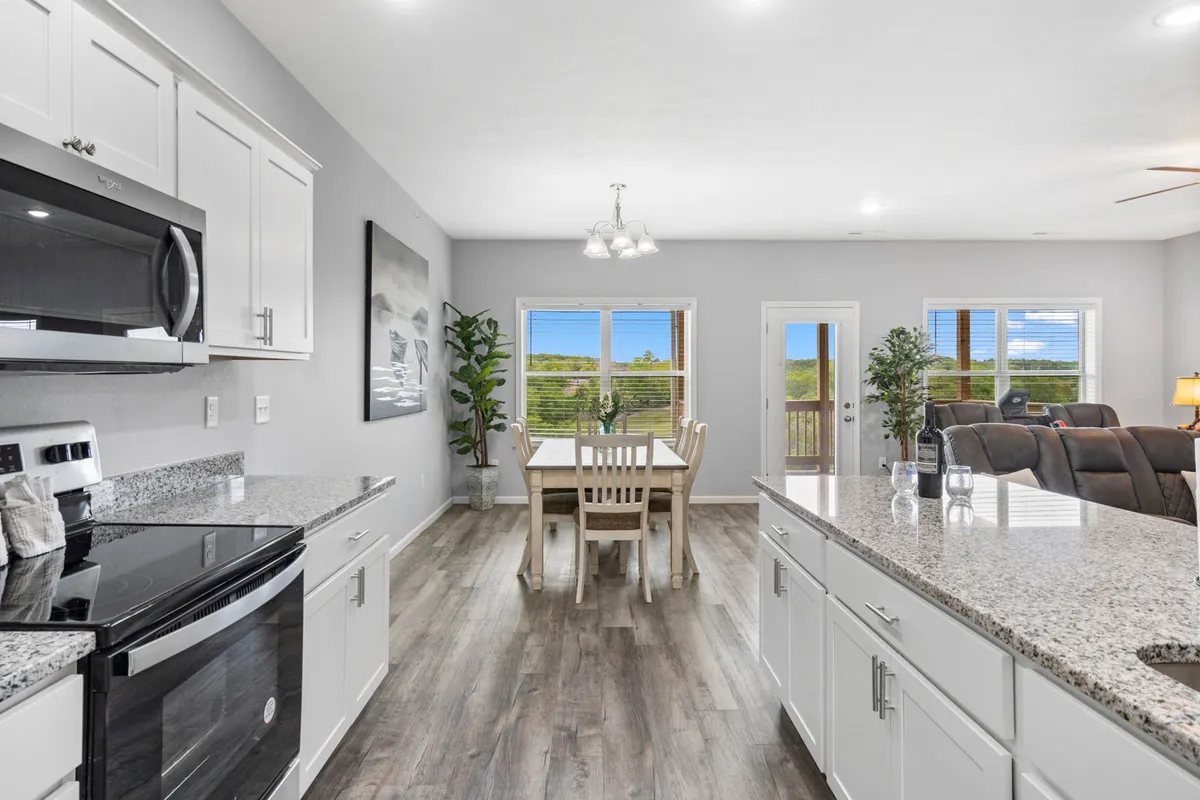 A bright interior scene showing a granite kitchen countertop, dining table, large windows letting in sunlight, a green plant, and comfortable seating. The overall mood is welcoming, ideal for family meals or casual gatherings in a modern setting.