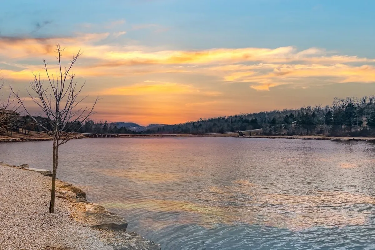 A tranquil lakeside scene featuring a calm lake at sunset, with a tree framing the shoreline and the serene water reflecting the colorful sky. The natural beauty and peacefulness of the area are emphasized, connecting visitors to the outdoor adventure and lakeside relaxation mentioned in the section.