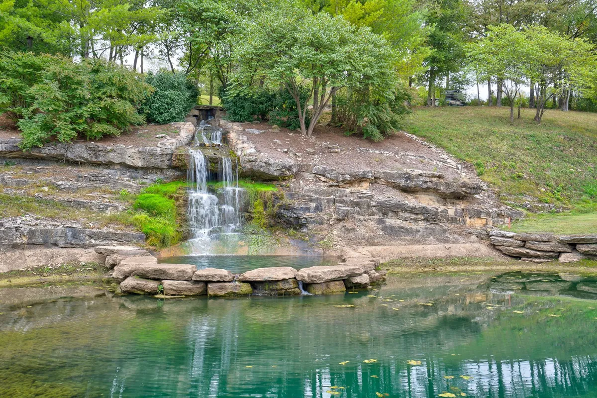 A tranquil reflective pond framed by lush greenery and a gentle waterfall, with natural rock formations. The area looks ideal for leisurely walks, peaceful moments by the water, and the enjoyment of nearby nature trails.