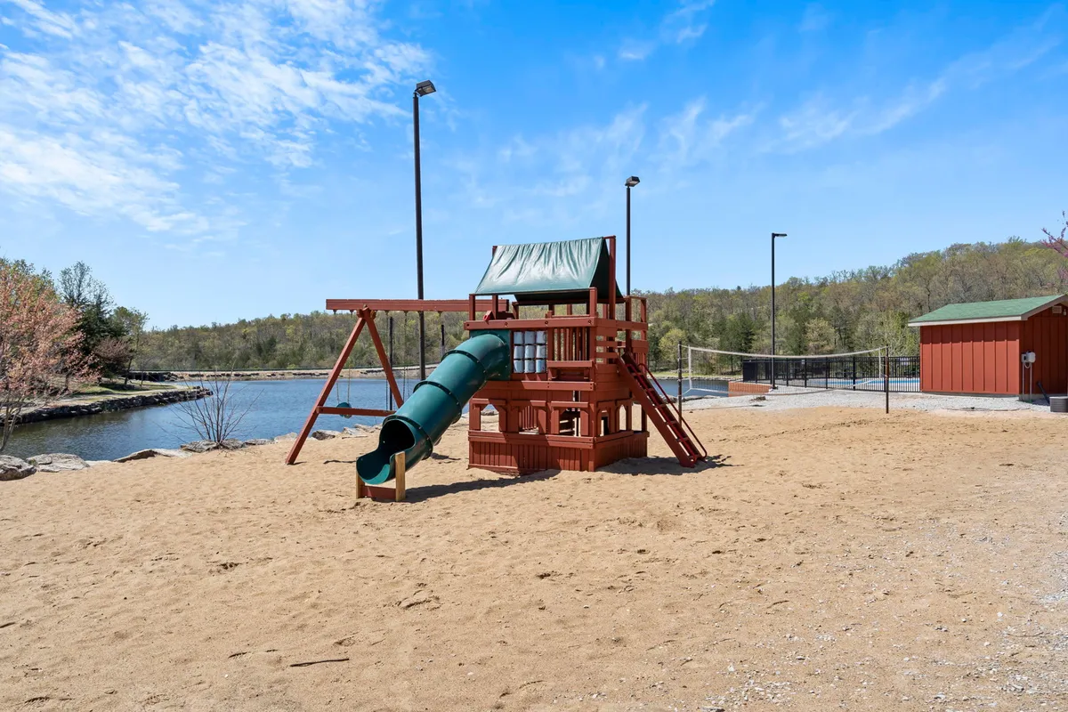 A vibrant playground scene set beside a serene water body, surrounded by greenery. Children are seen enjoying the play structures, slides, and sandbox, while families gather for picnics or outdoor games, filling the air with laughter and joy.