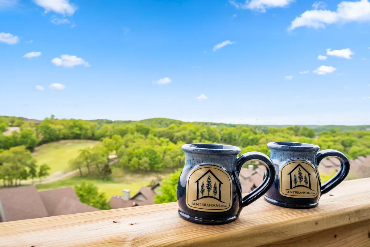 Two coffee mugs rest on a ledge or balcony with an expansive green landscape and blue sky in the distance, evoking a serene morning ritual. The composition draws attention to the peaceful connection with the outdoors.