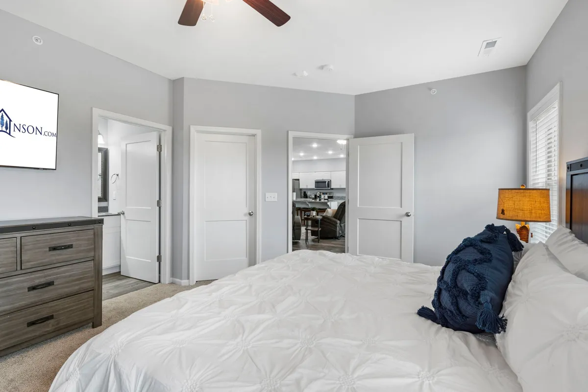 A serene bedroom with a neatly made bed adorned with decorative pillows, a dresser topped with a television, and a contemporary light fixture overhead. Sunlight filters softly through a nearby window, creating a peaceful, airy environment ideal for unwinding or enjoying a cozy movie night in bed.