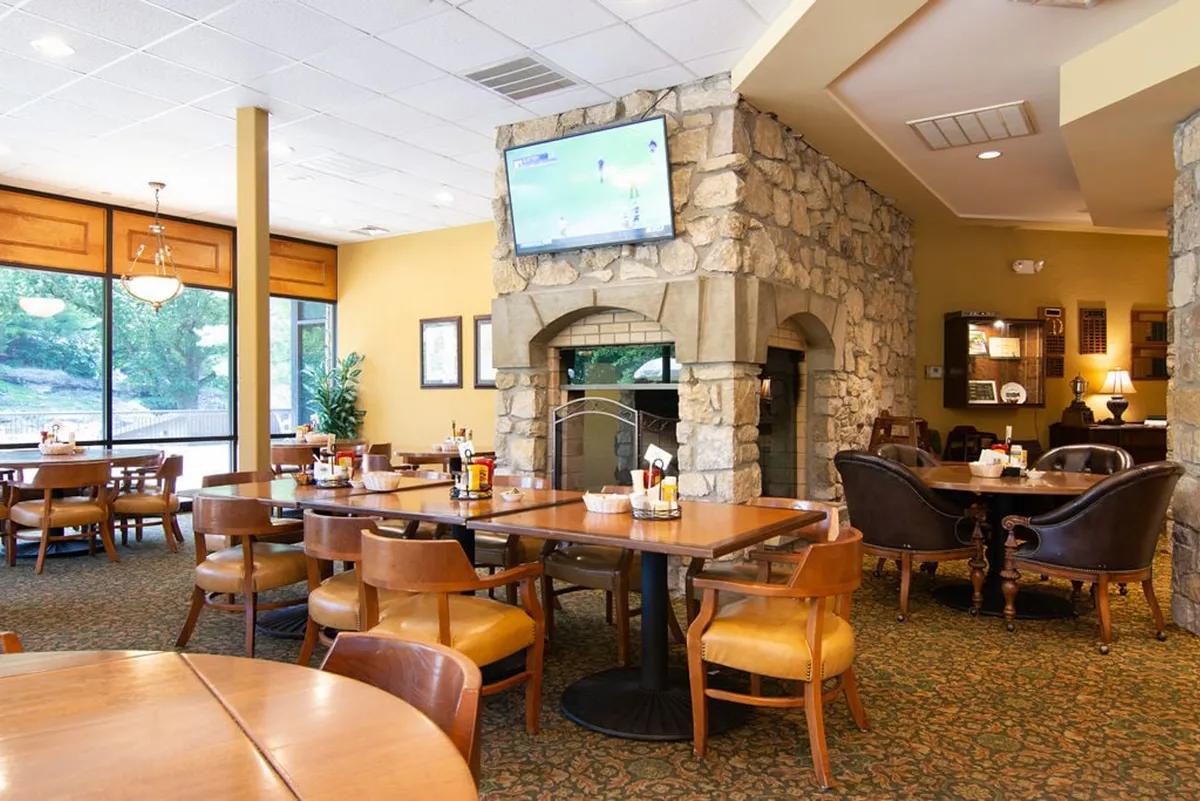 An inviting clubhouse interior featuring a cozy stone fireplace, large windows with views of the course, and dining tables arranged for socializing or relaxing after a round. A television and comfortable seating complete the welcoming atmosphere.