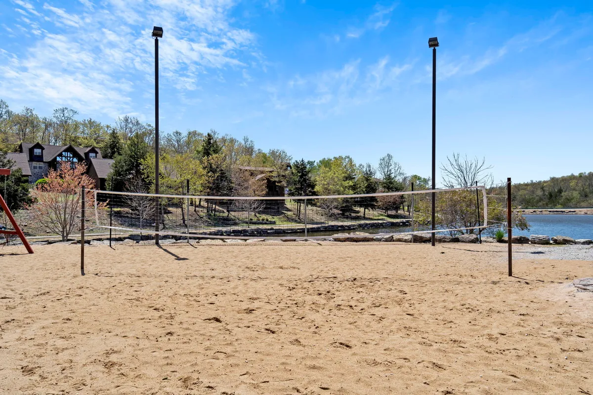 An energetic outdoor volleyball court with a sandy base located near the lake, surrounded by greenery and with a distant building visible. The space is alive with potential for games, group activities, and social gatherings by the water.