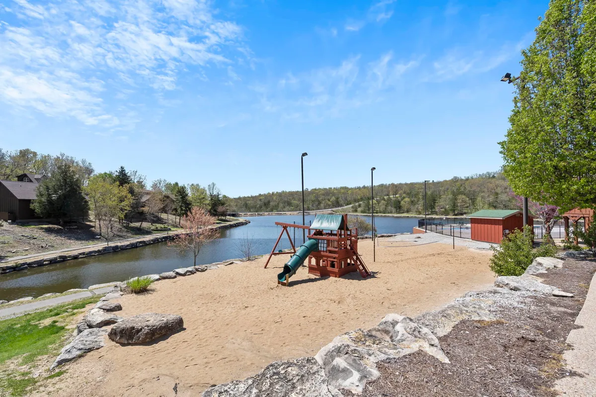 A lively scene featuring a playground set on sandy ground adjacent to a tranquil water body, framed by lush trees. Children can be seen playing, while families gather for picnics or leisurely moments by the water's edge.
